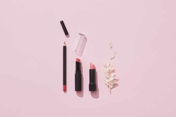 Flat lay of lipsticks and lip pencil with leaves on pink background.